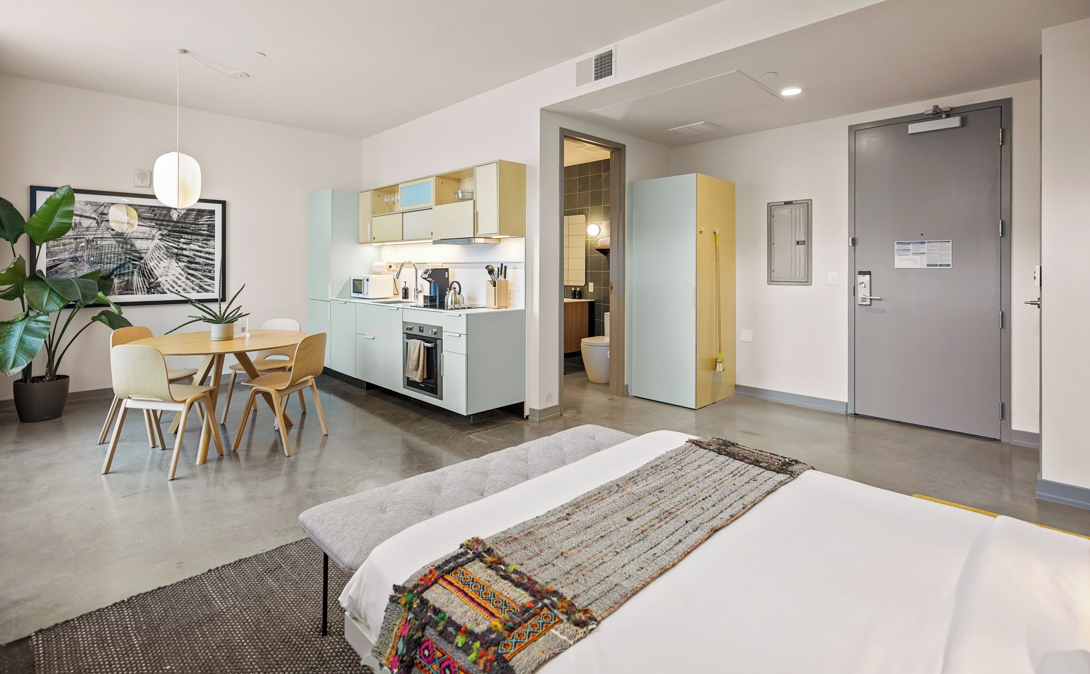 A modern hotel room with a bed, a dining table with chairs, a kitchen area, and a bathroom.