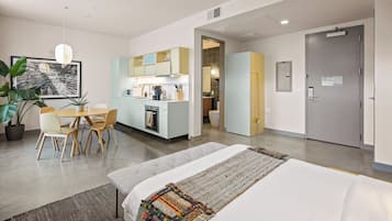 A modern hotel room with a bed, a dining table with chairs, a kitchen area, and a bathroom.