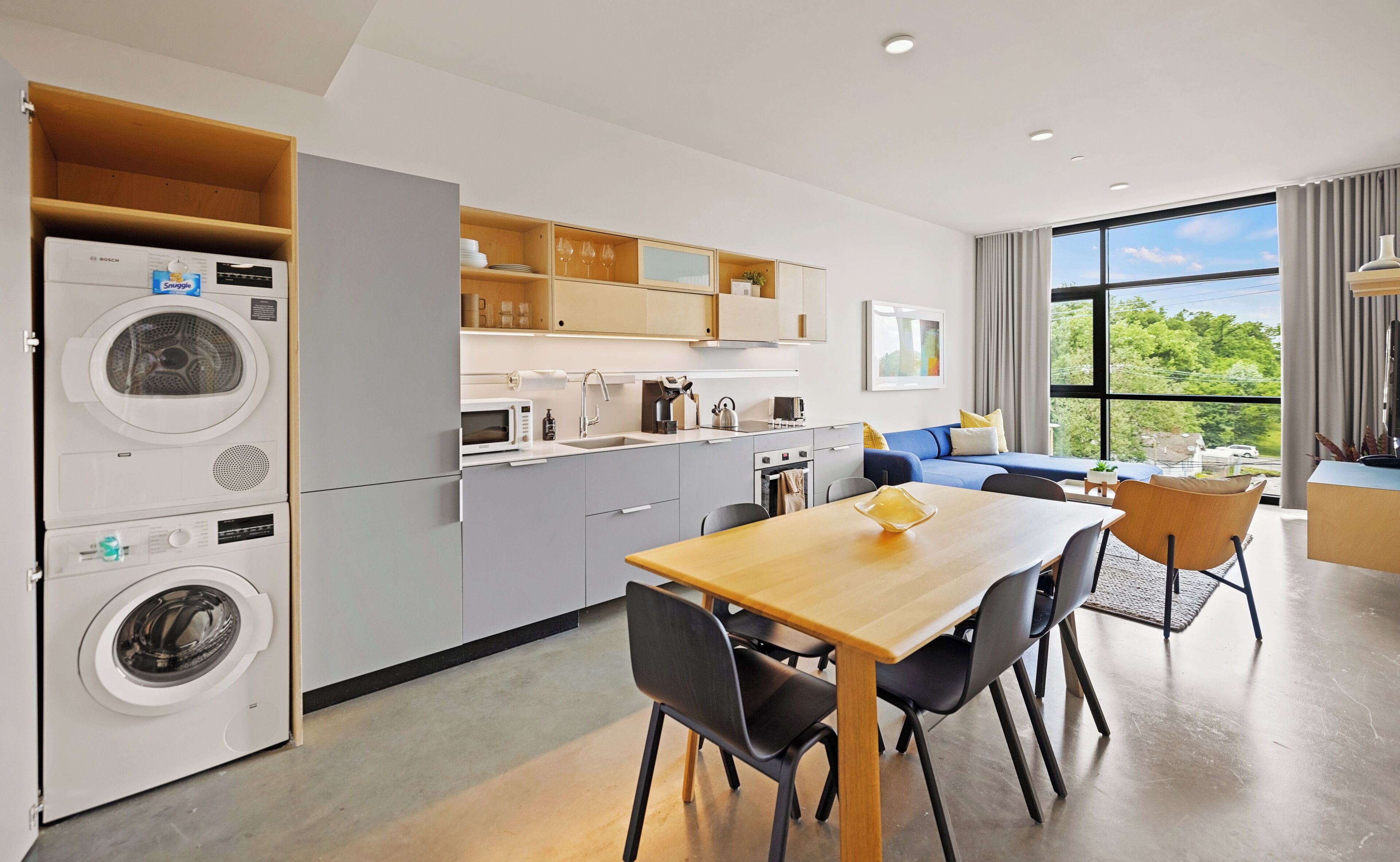 Une cuisine moderne dotée d’appareils électroménagers intégrés, un coin repas avec une table et des chaises en bois, et une grande fenêtre offrant une vue sur la verdure.