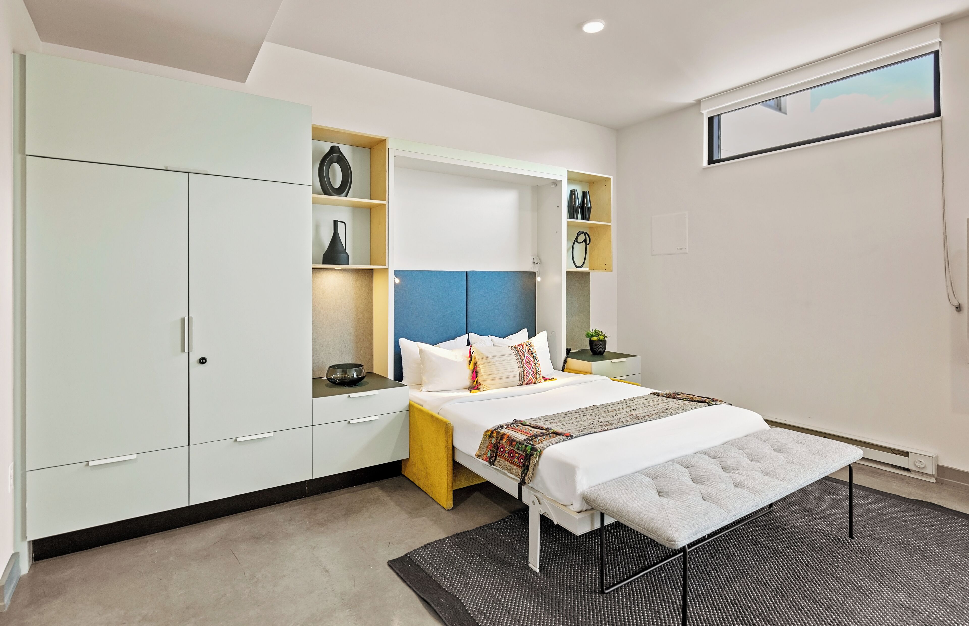 A modern bedroom with a bed, built-in storage, a bench, and a window.