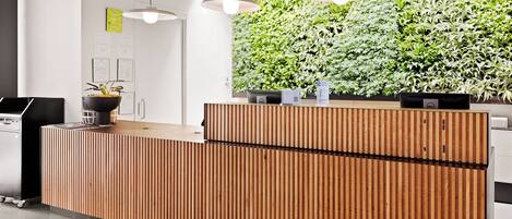 Un mostrador de recepción con detalles de madera, una pared verde con plantas e iluminación colgante.