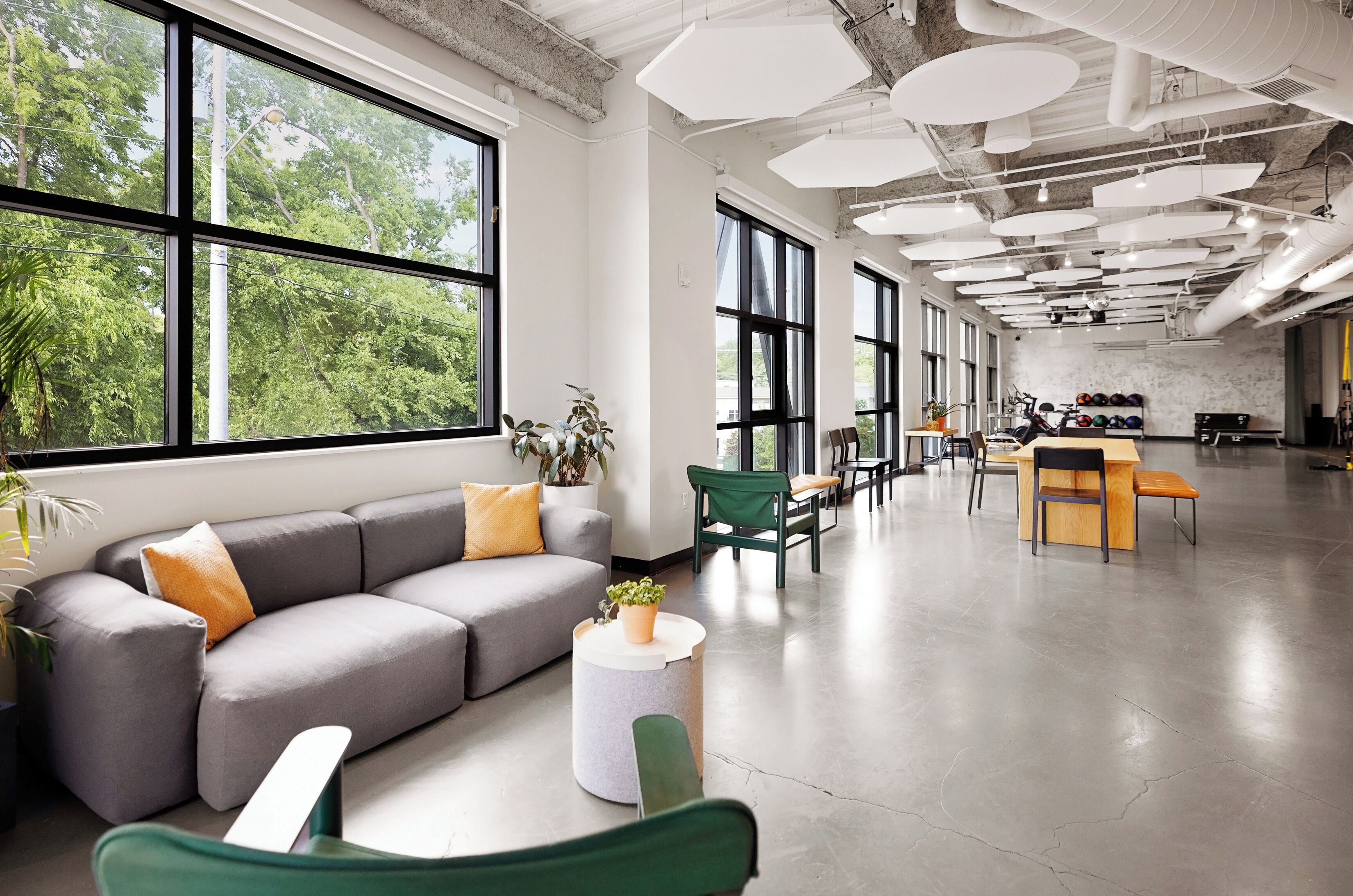 A spacious room with a grey sofa, green chairs, a round table, and large windows offering a view of trees.