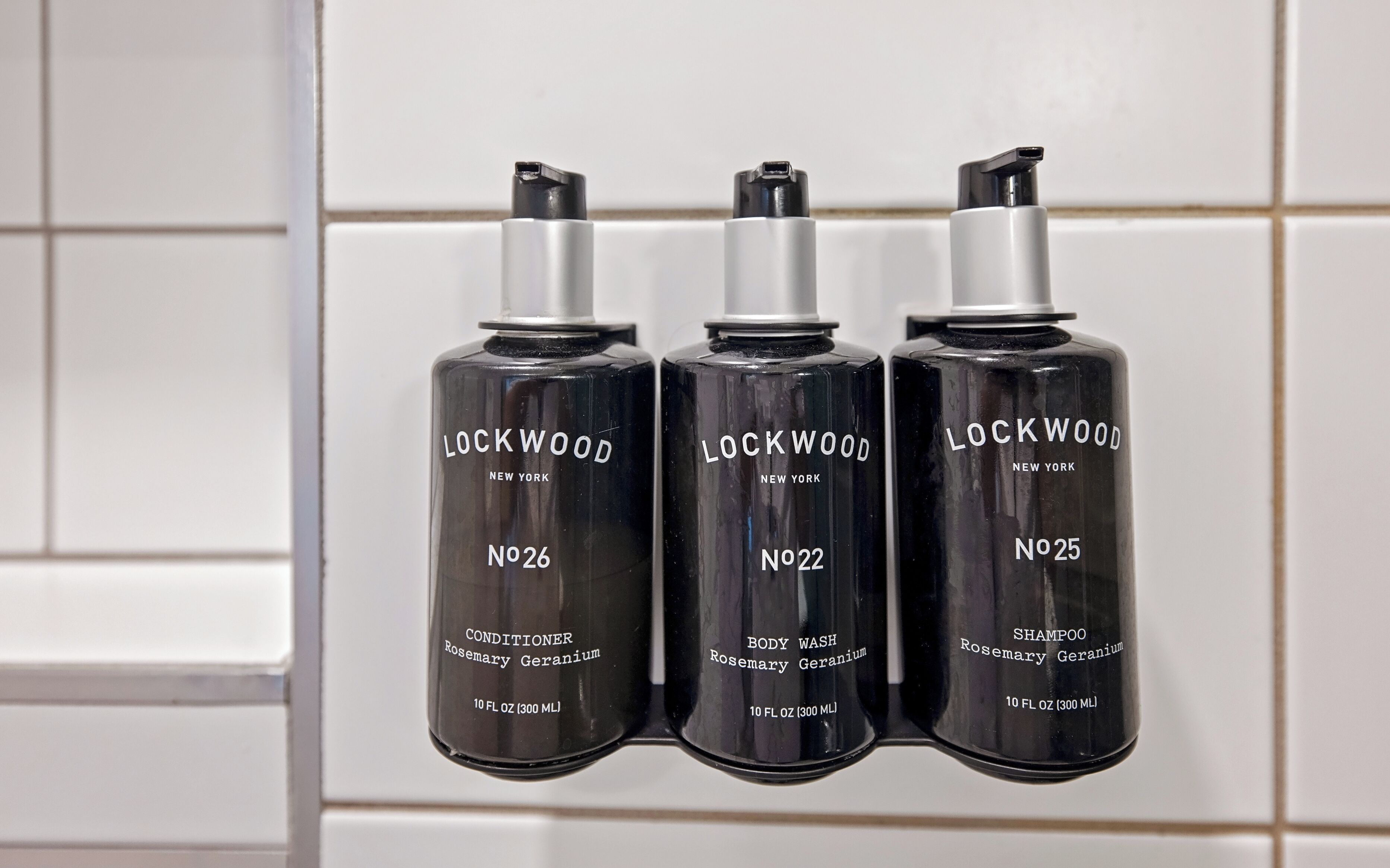 Three Lockwood New York products: No. 26 Conditioner, No. 22 Body Wash, and No. 25 Shampoo, all in black bottles with silver caps, displayed against a tiled wall.