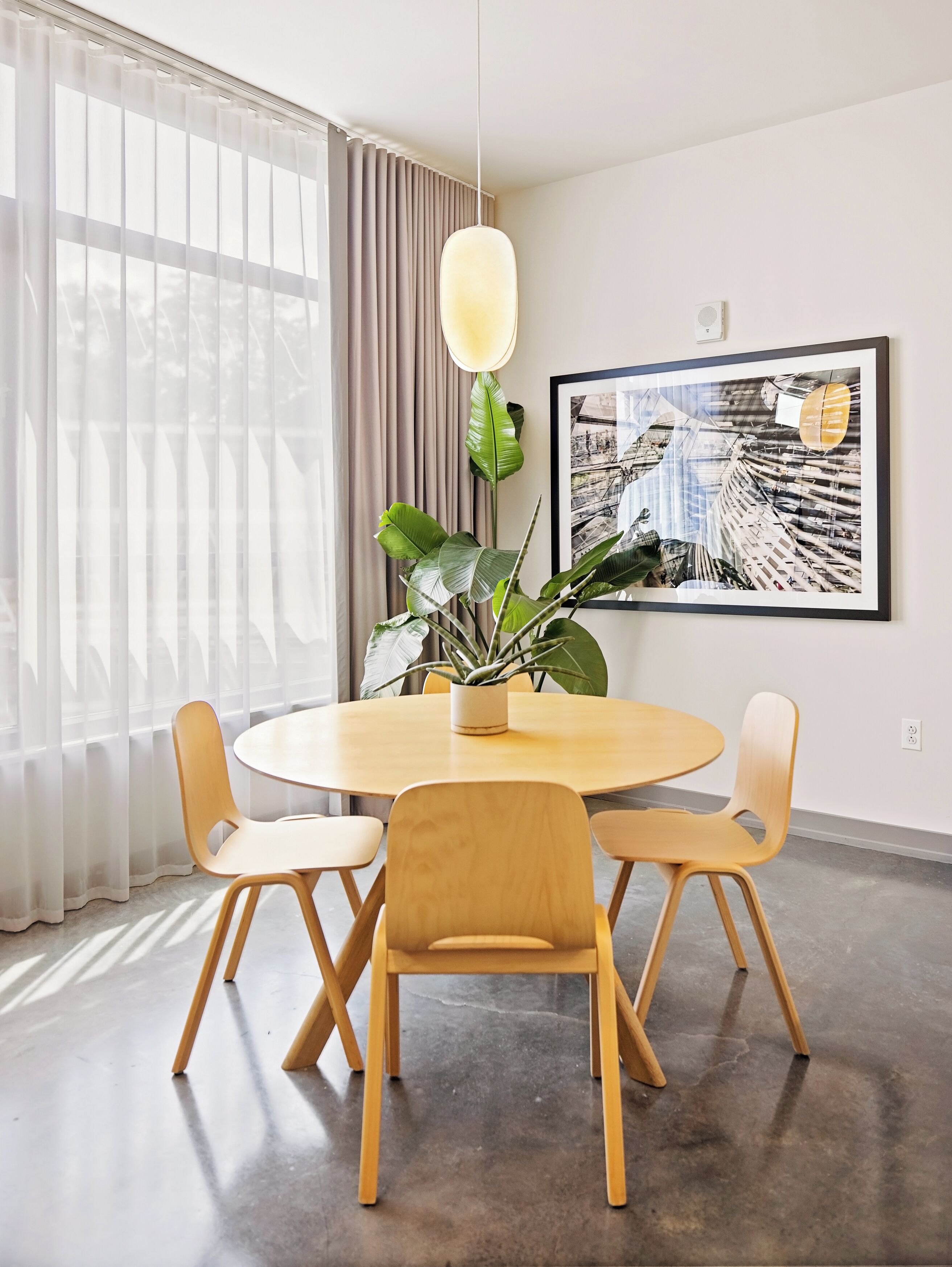 A modern dining area with a round table, four wooden chairs, a potted plant, a hanging light fixture, and a framed photograph on the wall.
