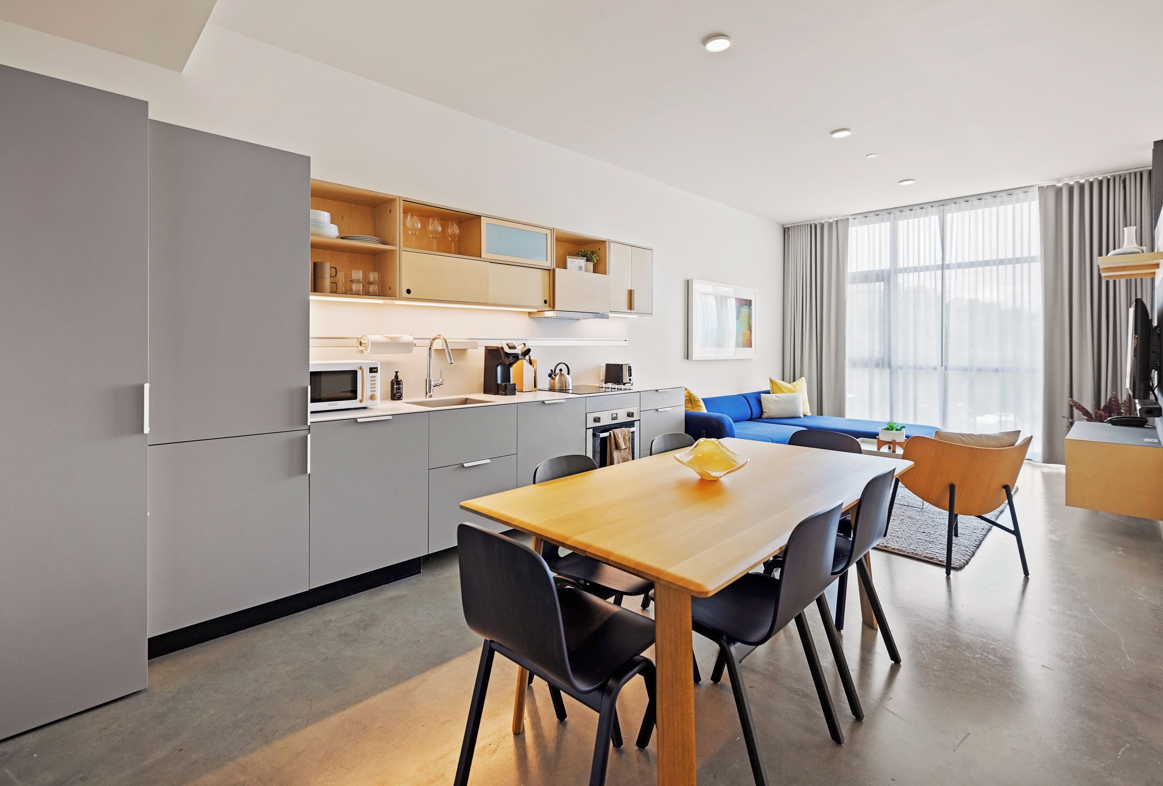 A modern kitchen with grey cabinets, a wooden dining table, and a blue sofa in the living area.