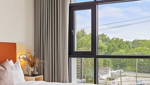 A bedroom with a large window, a bed, a nightstand, and a view of a street. - Harthall Hotel (Nashville)