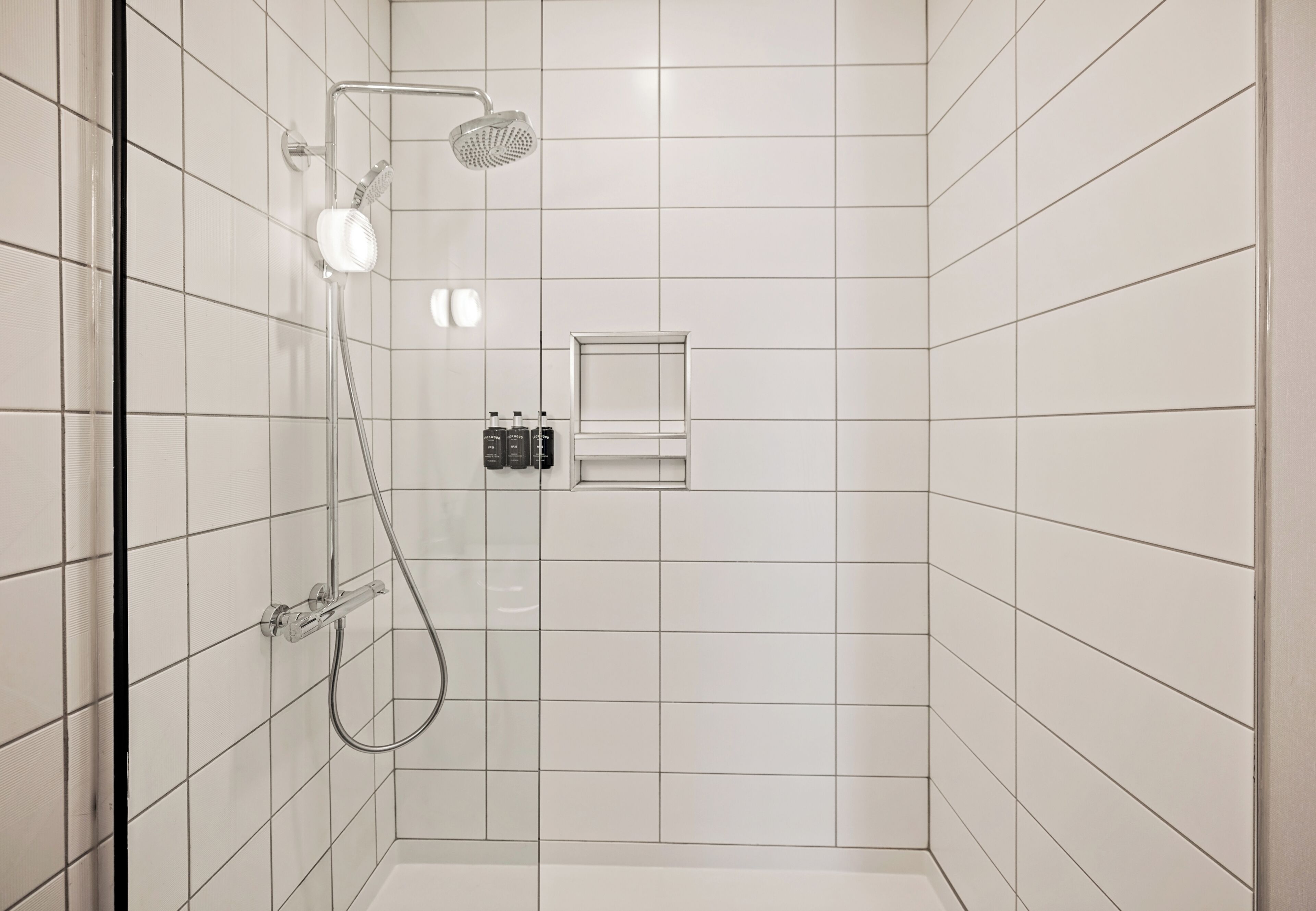 Eine Dusche mit Duschbrause und fest installierter Brause, eine kleine Ablage mit Pflegeprodukten sowie eine Glastür.