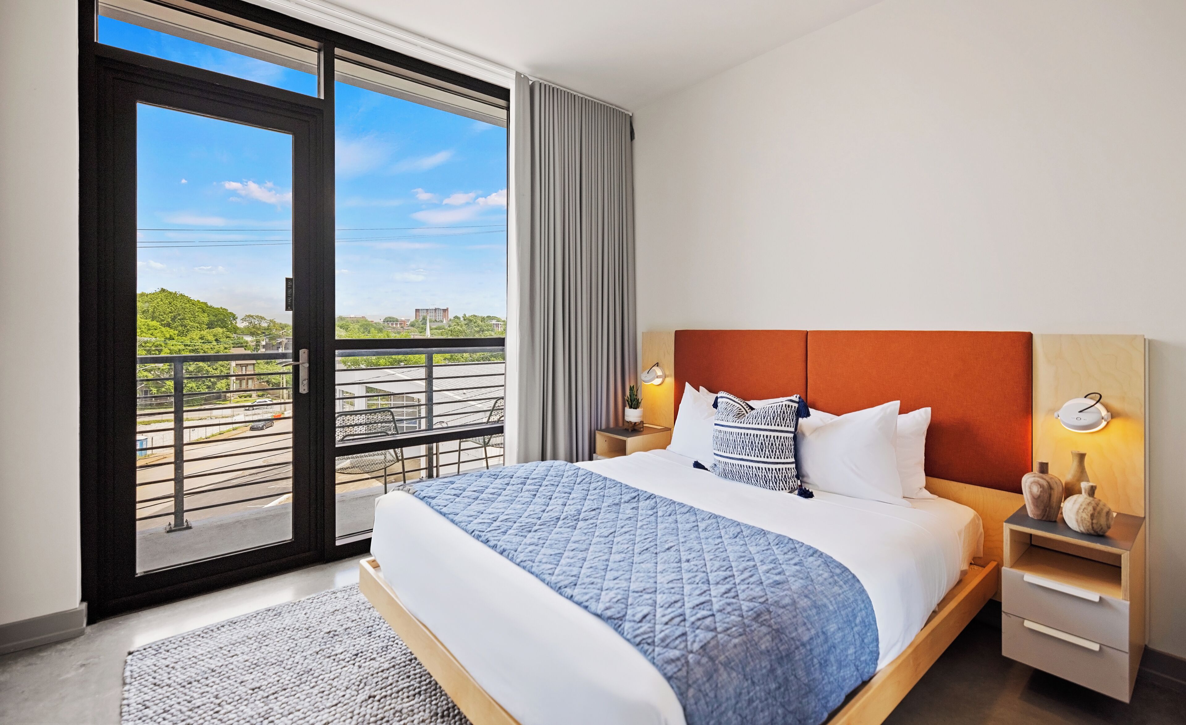 Hotelkamer met een groot bed, nachtkastje, een wandlamp en uitzicht op een groen landschap door een schuifdeur.