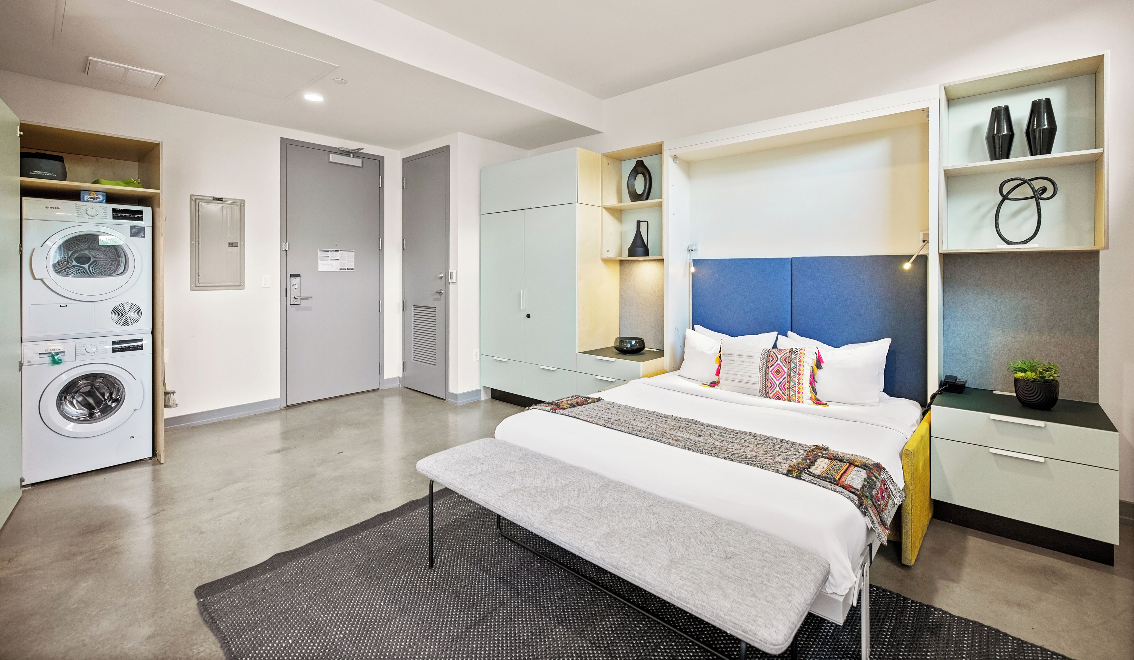 A modern hotel room with a bed, built-in wardrobe, a washing machine, and a small shelf with decorative items.