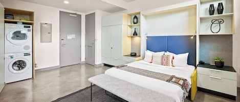 Una habitación de hotel moderna con cama, armario empotrado, lavadora y un pequeño estante con objetos decorativos.