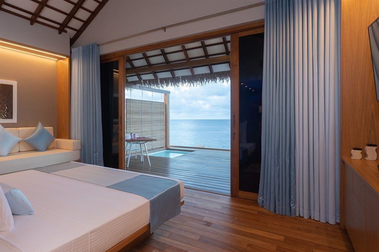A bedroom with a bed, a sofa, a small table, and a view of the sea through a sliding door.