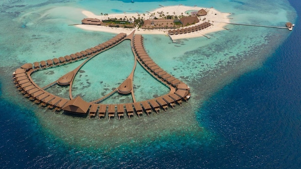 Vue aérienne d’un complexe hôtelier avec des bungalows sur pilotis reliés par un pont, des eaux turquoise cristallines et une plage de sable.
