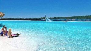 A beach scene with clear turquoise water, a sandy shore, and a distant boat. - 5 room TampaGolfVillas by AmericanVacationLiving (Wesley Chapel)