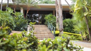 A staircase leading to a building with a glass facade, surrounded by lush greenery and palm trees.