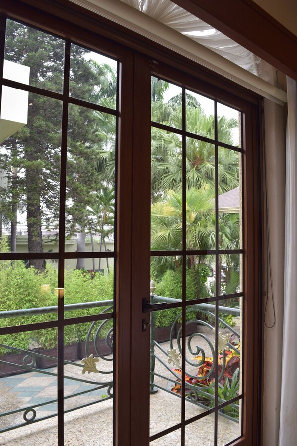 A view through a window with a grid pattern, revealing outdoor greenery and a paved area with a decorative metal railing. - Luxva Hotel Boutique (Guayaquil)