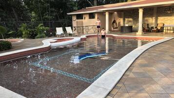 Una piscina con borde de azulejos, rodeada por un patio con sillas de descanso y un área de estar cubierta.
