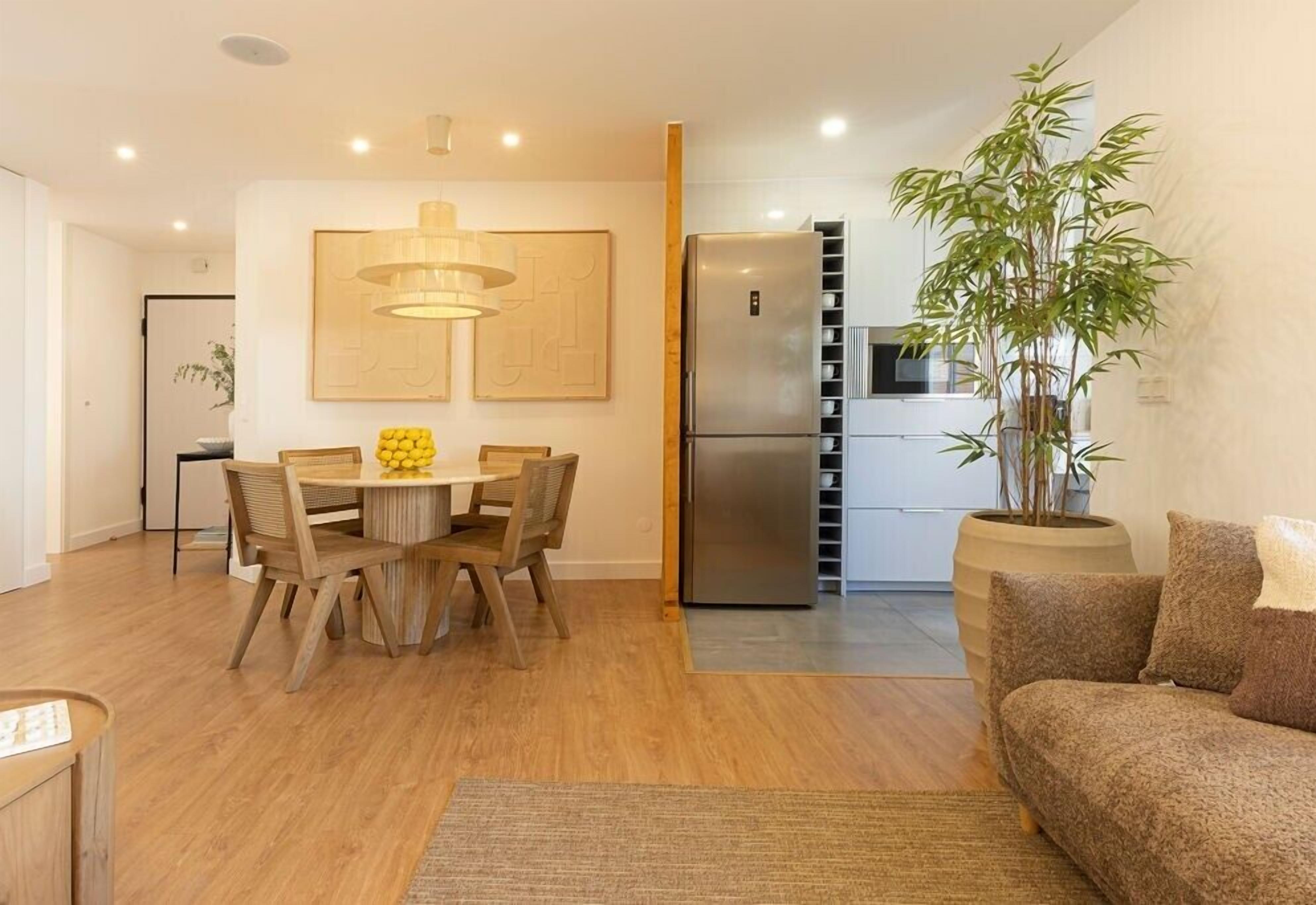 A modern kitchen with a built-in refrigerator, a dining area with a wooden table and chairs, and a living area with a sofa and a large potted plant.