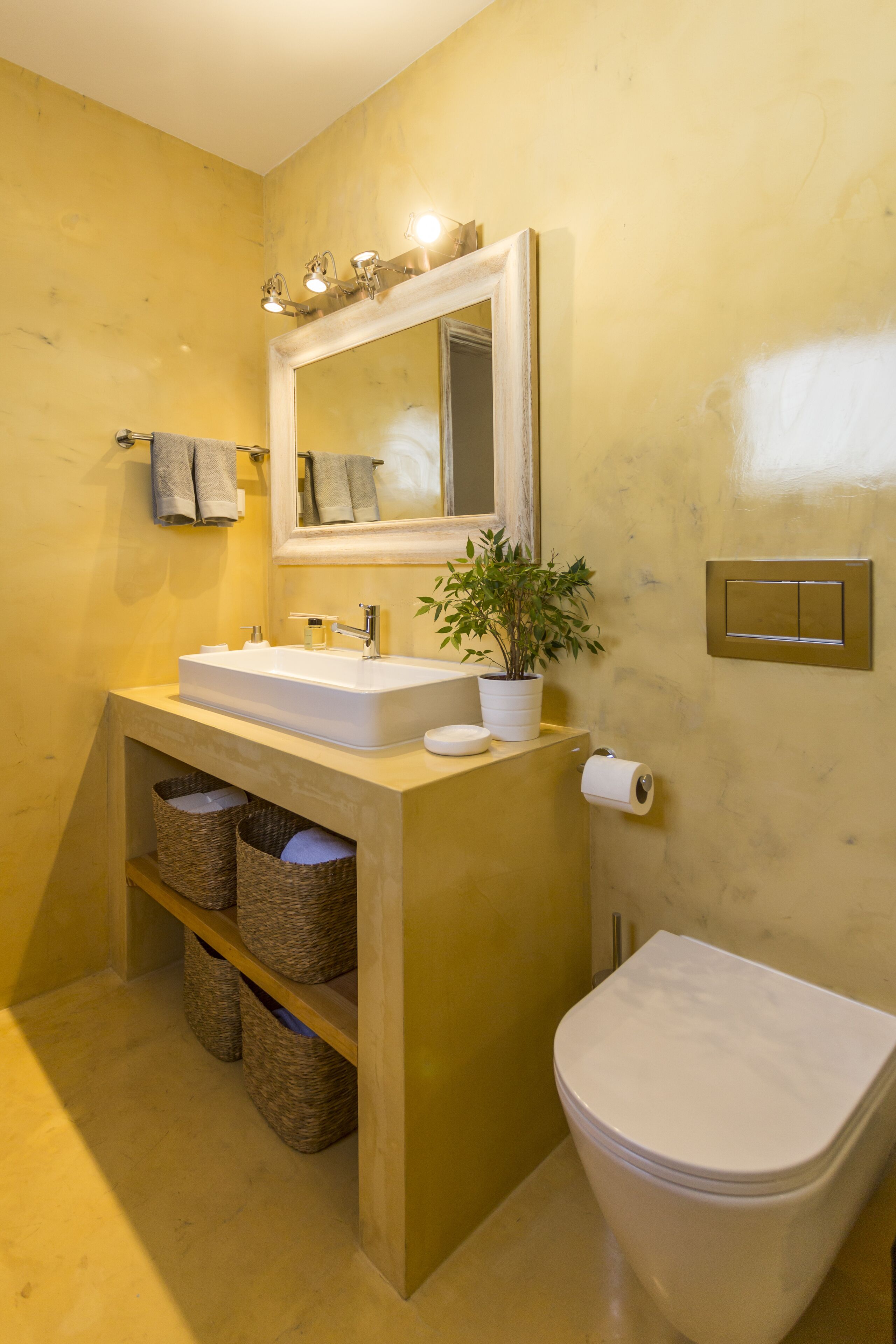 A bathroom with a white sink, mirror, and toilet. There are wicker baskets under the sink and a potted plant on the counter.