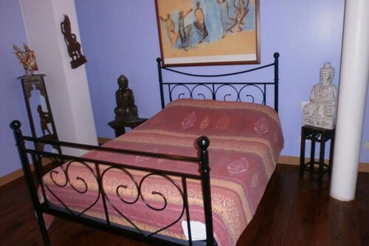 A room with a metal bed frame, a patterned bedspread, a painting on the wall, and a statue on a side table.