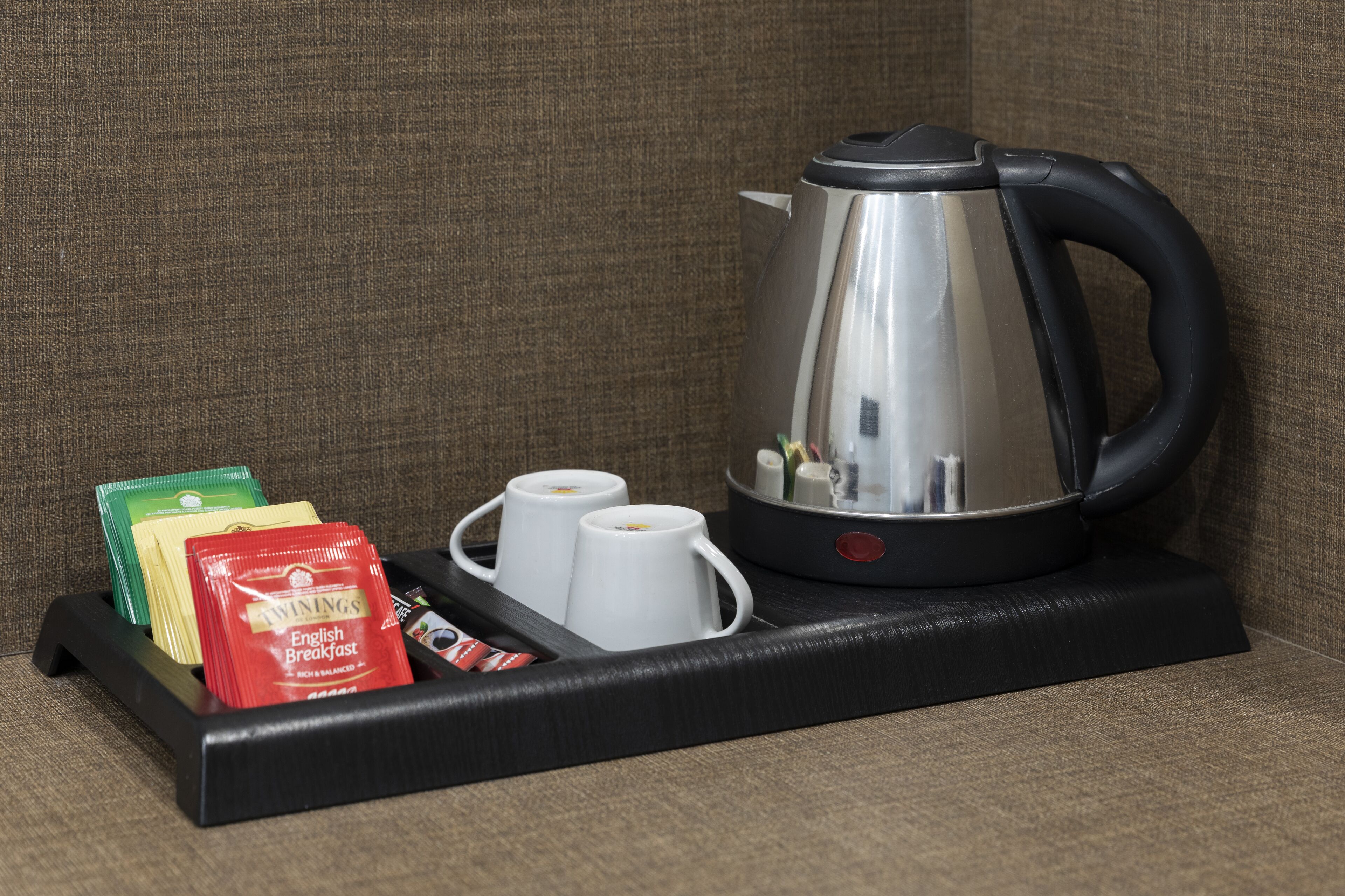Uma cafeteira com cabo preto e corpo de aço inoxidável sobre uma bandeja preta. A bandeja contém duas chávenas brancas e três saquetas de chá Twinings English Breakfast.