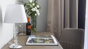 Habitación de hotel con una pequeña mesa, una lámpara, una botella de vino, un jarrón con flores y una silla.