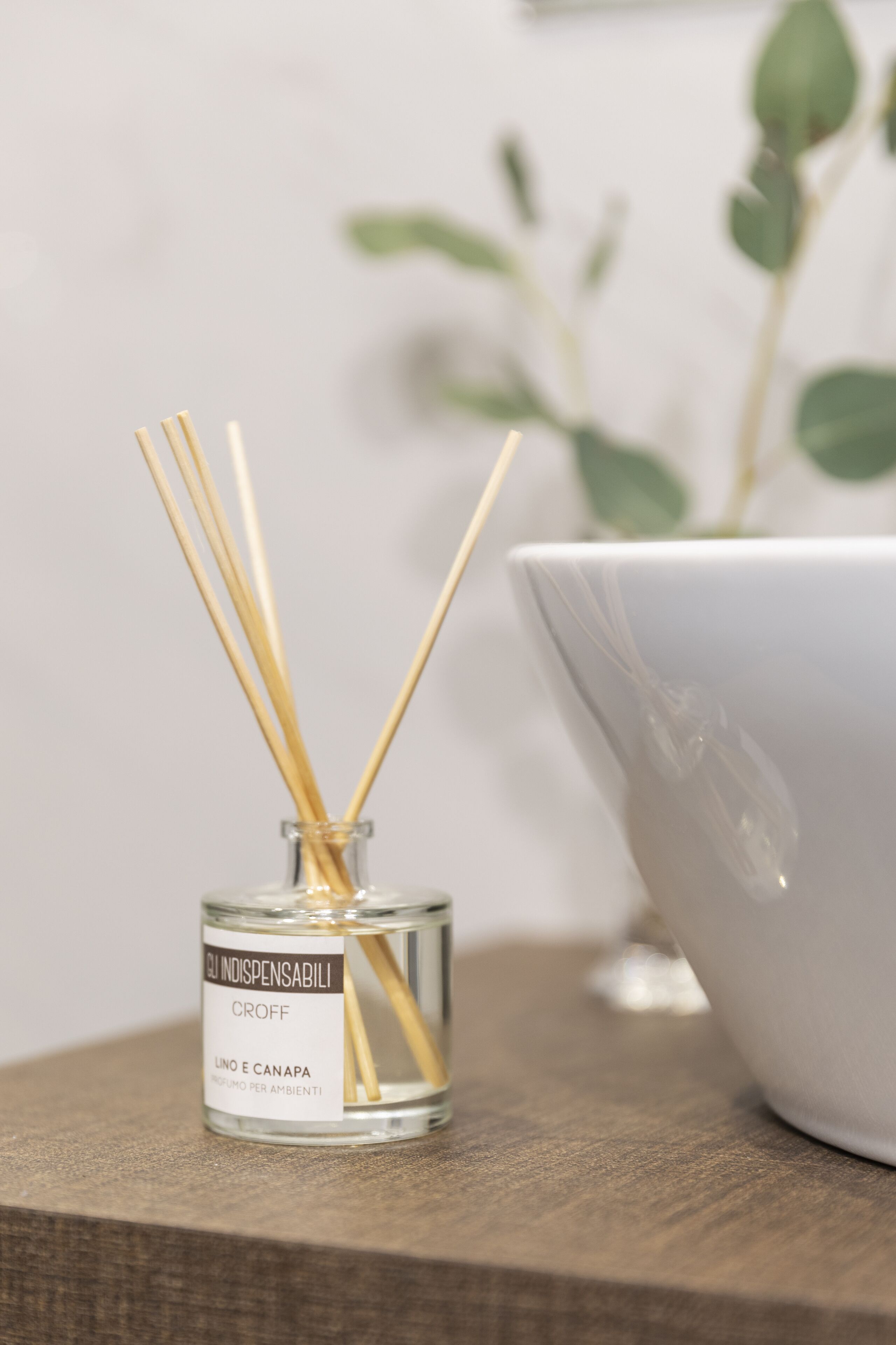 Pequeña botella de vidrio con palitos de junco y una etiqueta, sobre una superficie de madera, junto a un cuenco blanco.