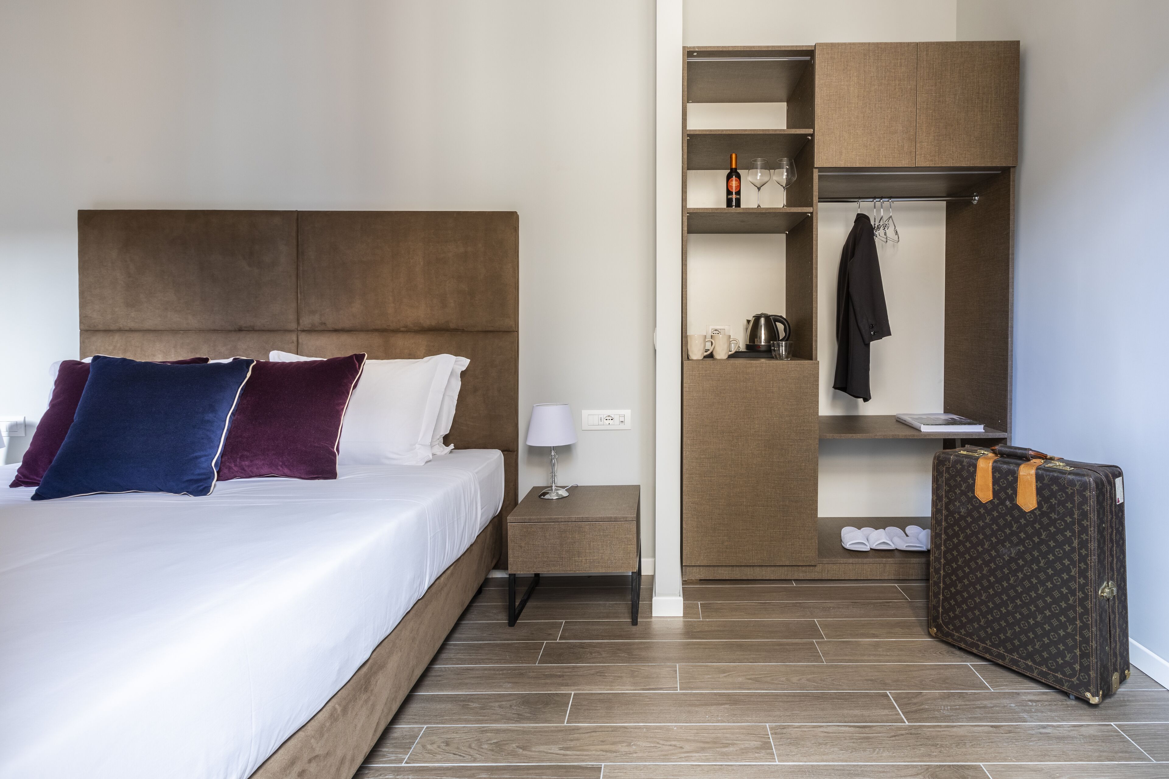 A hotel room with a bed, bedside table, lamp, and a wardrobe with a hanging coat.