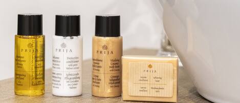 Three bottles of PRIJA products and a wooden soap bar on a wooden surface with a white bowl and green leaves in the background.