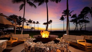 A stone fire pit surrounded by wicker furniture, set against a backdrop of palm trees and a sunset sky.