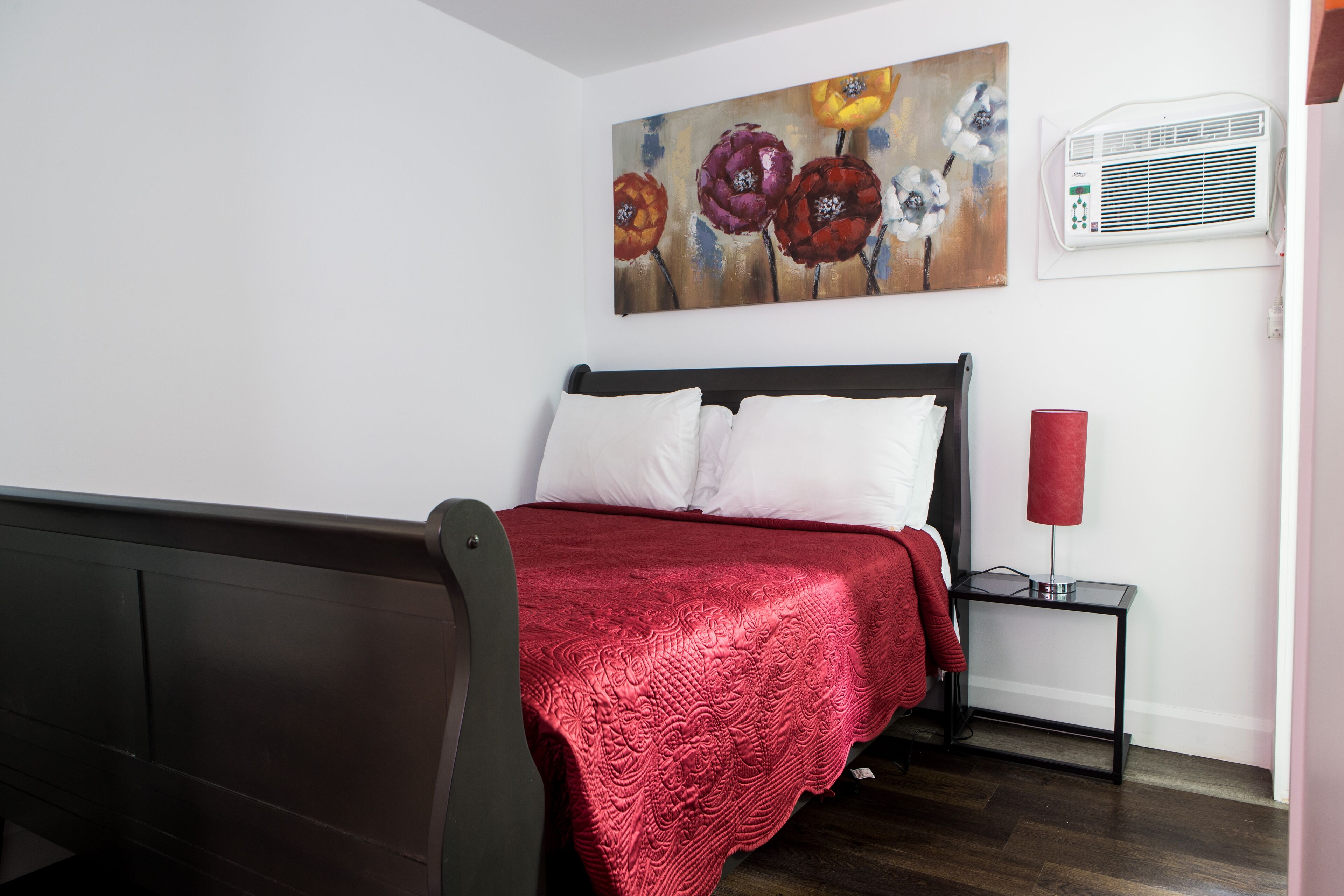 Un dormitorio con cama, una manta roja, una mesita de noche con lámpara y un cuadro en la pared.