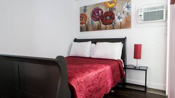 Un dormitorio con cama, una manta roja, una mesita de noche con lámpara y un cuadro en la pared.