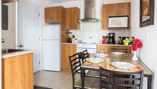 A kitchen with wooden cabinets, a white refrigerator, a stove, a microwave, and a dining table set with plates and a vase of flowers. - Auberge Motel La Référence (Trois-Rivieres)