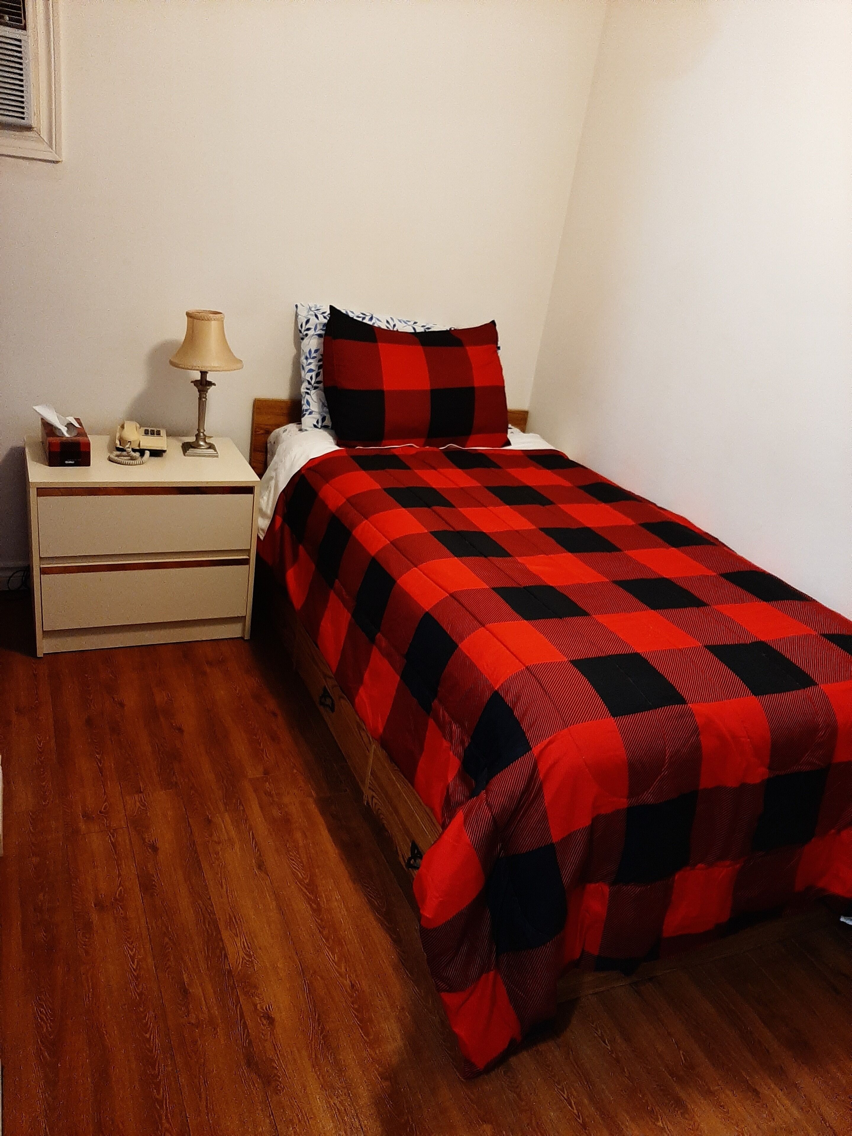 Een kleine, netjes ingerichte slaapkamer met een houten vloer, een eenpersoonsbed met een rood-zwarte geruite plaid, een witte nachtkastje met een lamp en een kussen met dessin.