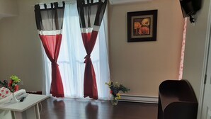 A room with a white desk, a telephone, and a vase with flowers. There are curtains with red and white panels, and a framed picture on the wall. - Auberge Motel La Référence (Trois-Rivieres)