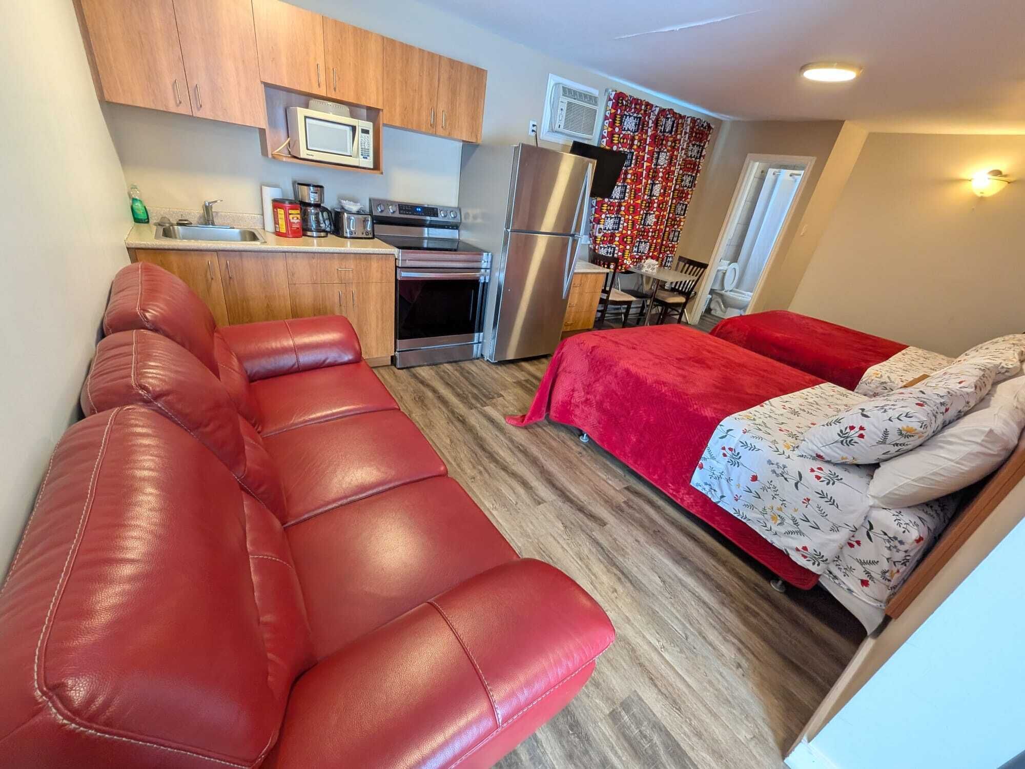 Un espacio habitable compacto con kitchenette, un sofá de cuero rojo, cama con ropa de cama floral y zona de comedor.