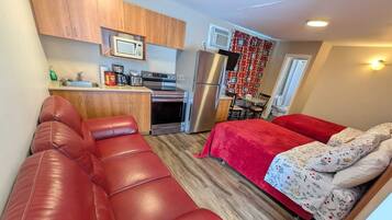 Un espacio habitable compacto con kitchenette, un sofá de cuero rojo, cama con ropa de cama floral y zona de comedor.