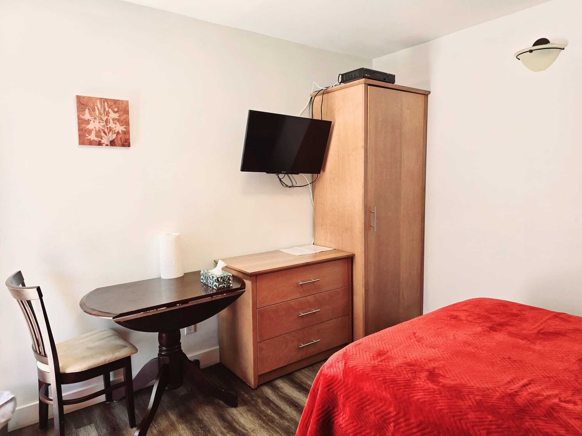 A small room with a wooden desk, a chair, a television mounted on the wall, a wardrobe, and a bed with a red blanket.