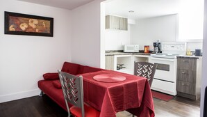 A compact kitchen with white appliances, a dining area with a red table and matching chairs, and a framed artwork on the wall. - Auberge Motel La Référence (Trois-Rivieres)