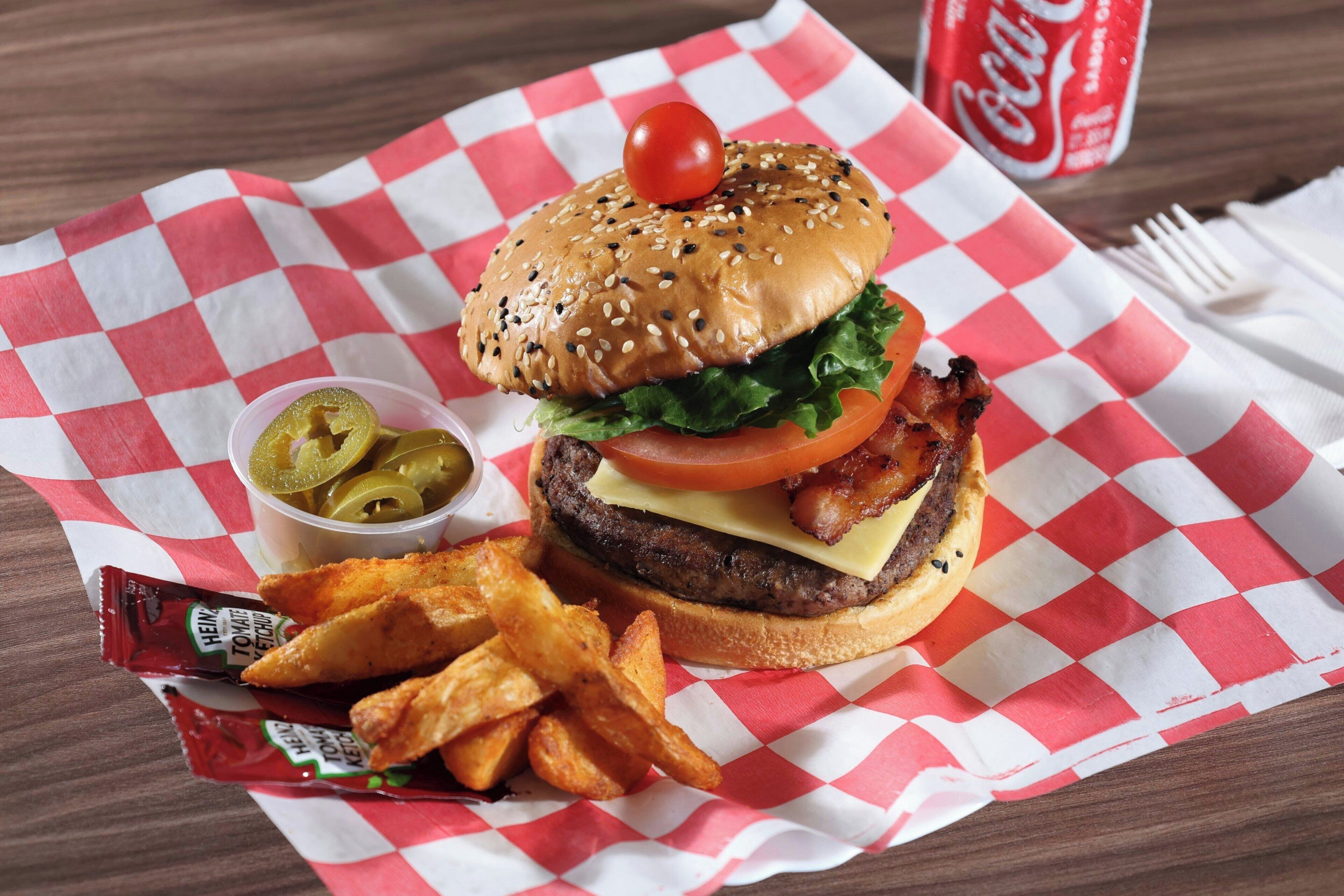 A burger with lettuce, tomato, and bacon on a sesame seed bun, served with fries and pickles, on a checkered paper.