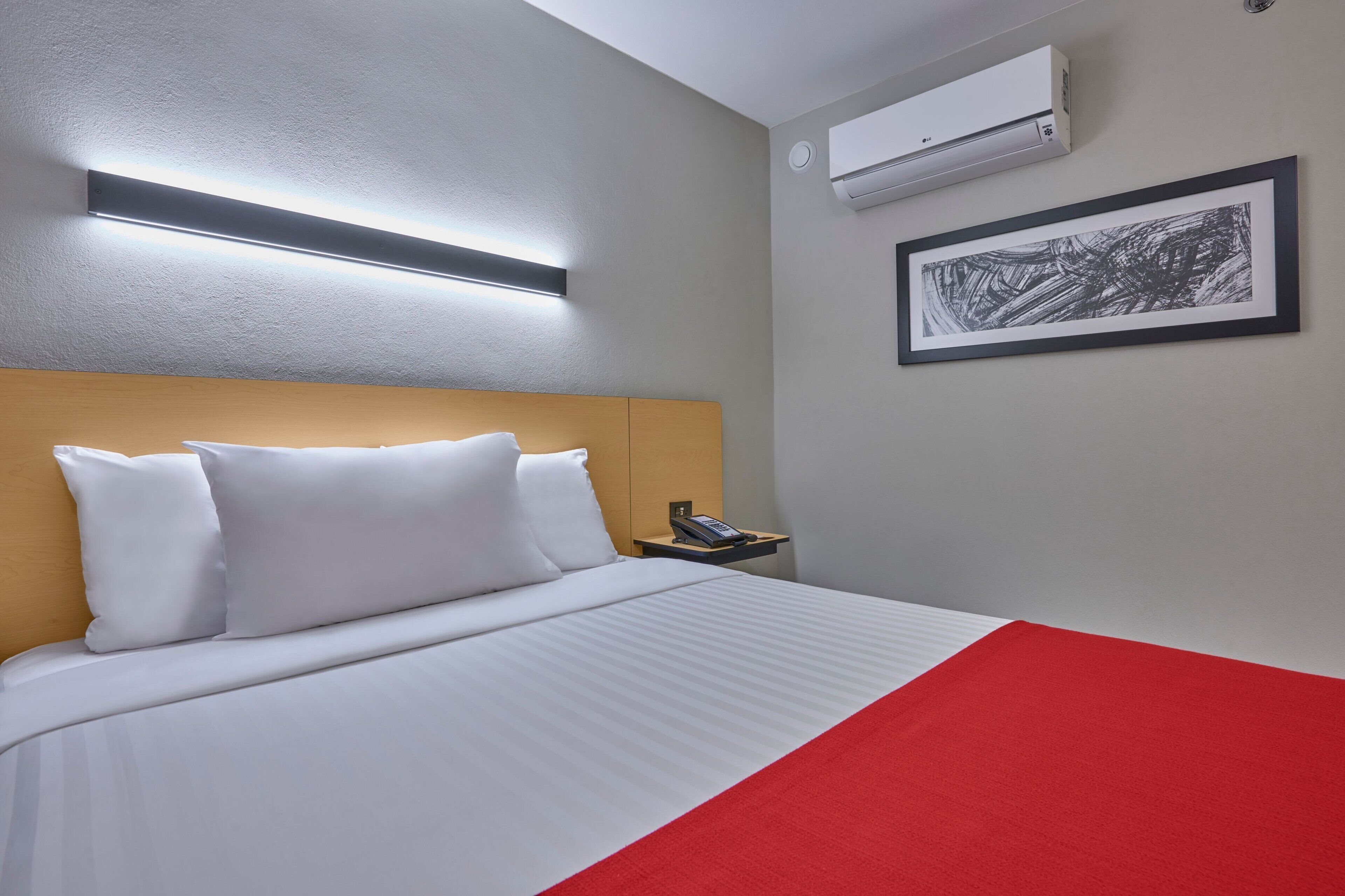 A hotel room with a bed, a bedside table with a phone, an air conditioner, and a framed artwork on the wall.