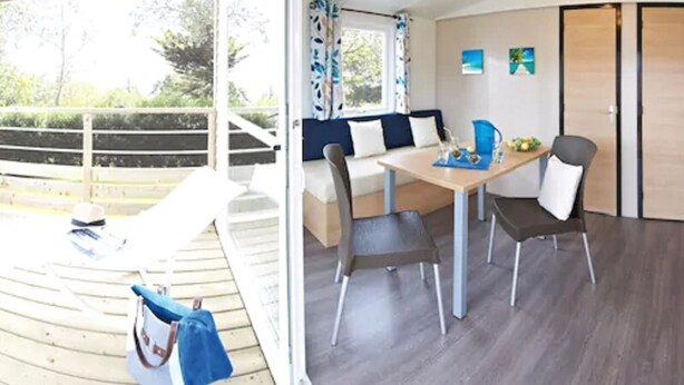 A modern room with a wooden deck, a sofa, a dining table with chairs, and a view of greenery outside. - Camping Haliotis (Pontorson)