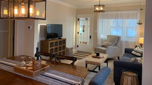 A spacious living room with a wooden coffee table, a sofa, and a TV. There is a dining area with a wooden table and chairs. The room has a large window with curtains and a chandelier. - On the Trail-Cedar Sauna & Close to Ski & Trails (Ironwood)