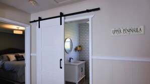 A bedroom with a bed, a sliding door leading to a bathroom with a sink and mirror, and a sign on the wall reading "Upper Peninsula." - On the Trail-Cedar Sauna & Close to Ski & Trails (Ironwood)