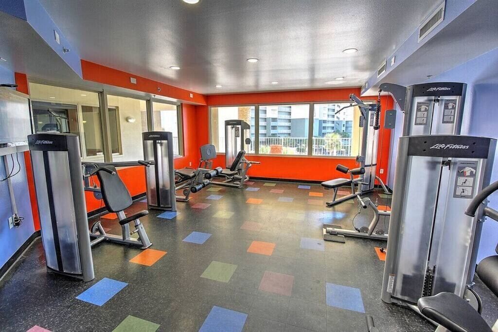 A gym with various exercise machines, a mirrored wall, and large windows with a city view.