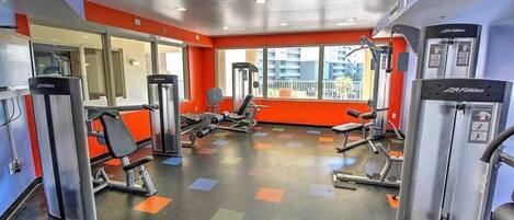 A gym with various exercise machines, a mirrored wall, and large windows with a city view.