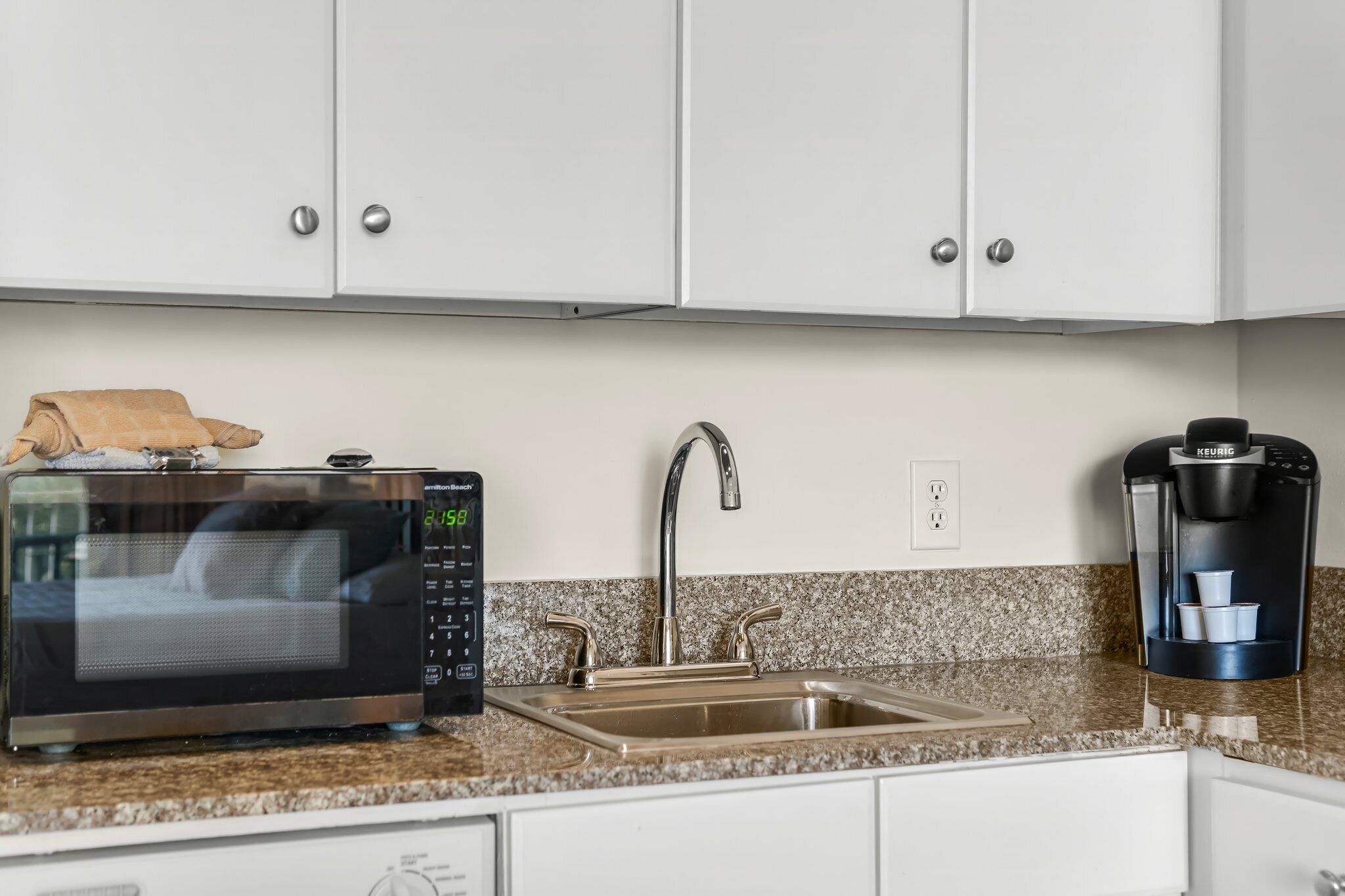Cozinha com armários brancos, pia de aço inoxidável, micro-ondas e cafeteira Keurig.