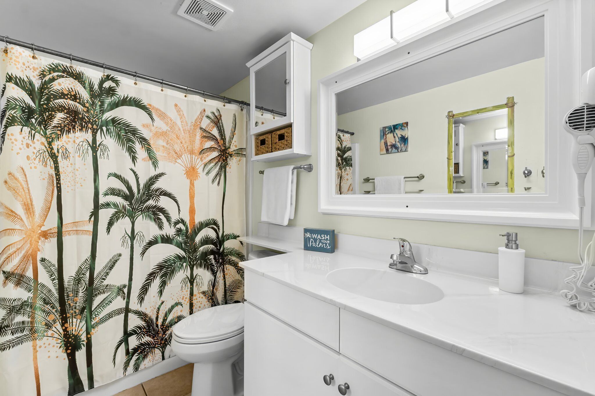 A bathroom with a white vanity, a large mirror, a towel rack, and a shower curtain with a palm tree design.