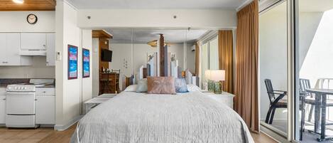 A modern hotel room with a large bed, a kitchenette, a dining area, and a balcony with curtains.
