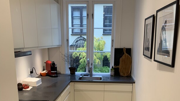 A compact kitchen with a black countertop, white cabinets, a built-in oven, and a window with a view of greenery and buildings. - Apartment 1 bedroom Grønnegade (Copenhagen)