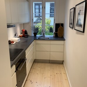 A compact kitchen with a black countertop, white cabinets, a built-in oven, and a window with a view of greenery and buildings. - Apartment 1 bedroom Grønnegade (Copenhagen)